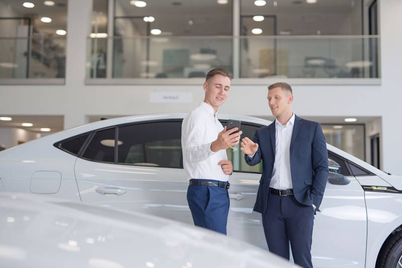 Praktikum im Vertrieb Zwei Männer im Autohaus zeigen auf ein Smartphone vor einem weißen Auto.